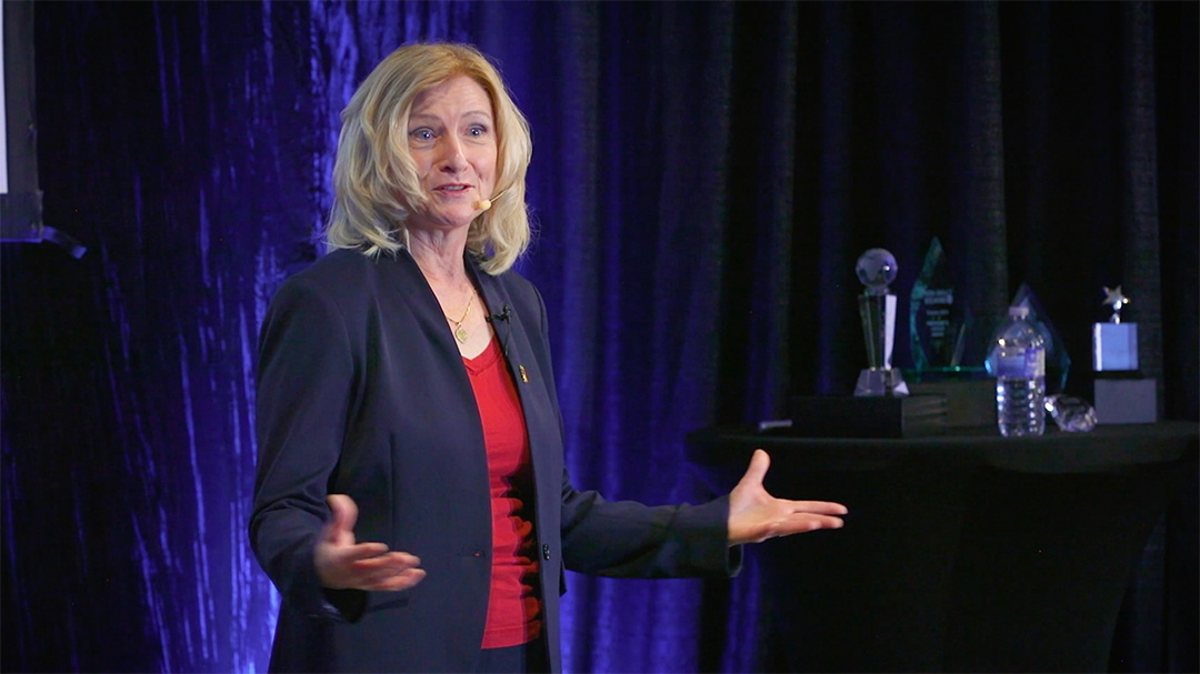 Iwona Buziak-Mohamed screenshot speaking. Dressed in a dark blue suit, red shirt, on a black and purple background.